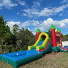 An inflatable bounce house and slide combo featuring bright green, yellow and red colors. The structure includes a large bounce area, a climbing wall, and a slide with a pool area at the bottom of the slide, with netted windows for visibility and airflow. Set up on a grassy lawn with trees in the background, the scene is lively and inviting, ideal for outdoor fun.