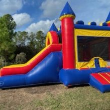 Bouncy castle with a large slide out the front of the bounce castle. There is a small slide out the side of the bounce castle. Bounce castle is red, blue and yellow.